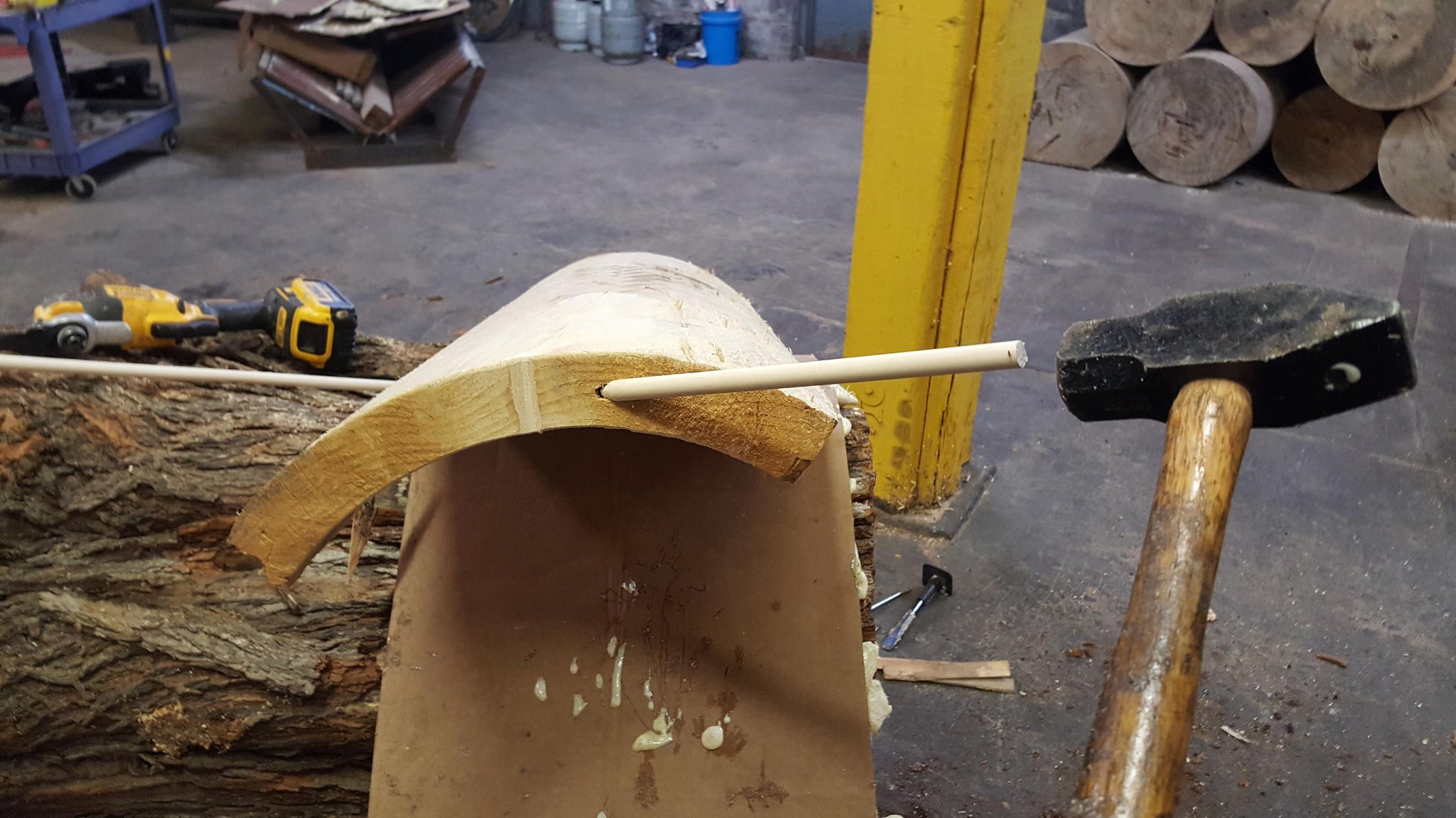 Dowel rod being inserted through a drilled channel spanning the crack from the top of the IC WOOD half-log — hammer and DeWalt drill visible in background, cardboard backing inside the hollow